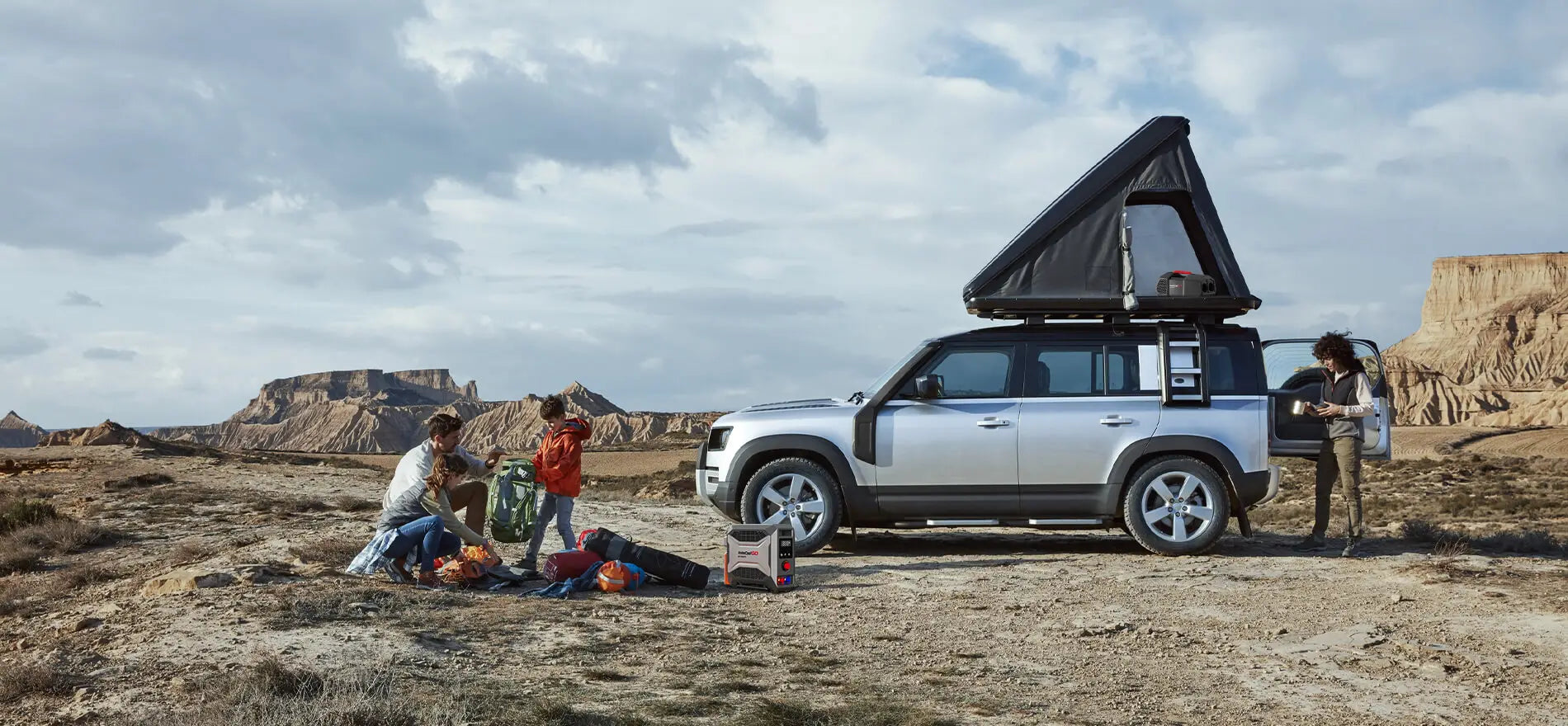 People camp in the wild with a silver SUV. The scene features the Fogatti InstaShower GO Outdoor Portable Water Heater, ready for outdoor use.