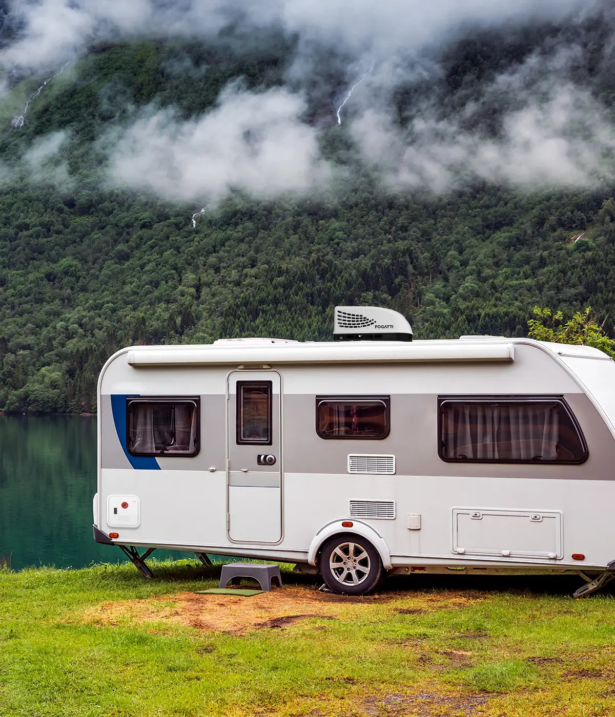A caravan by a tranquil lake, surrounded by lush mountains. The body of the caravan is equipped with a Fogatti - branded water heater.