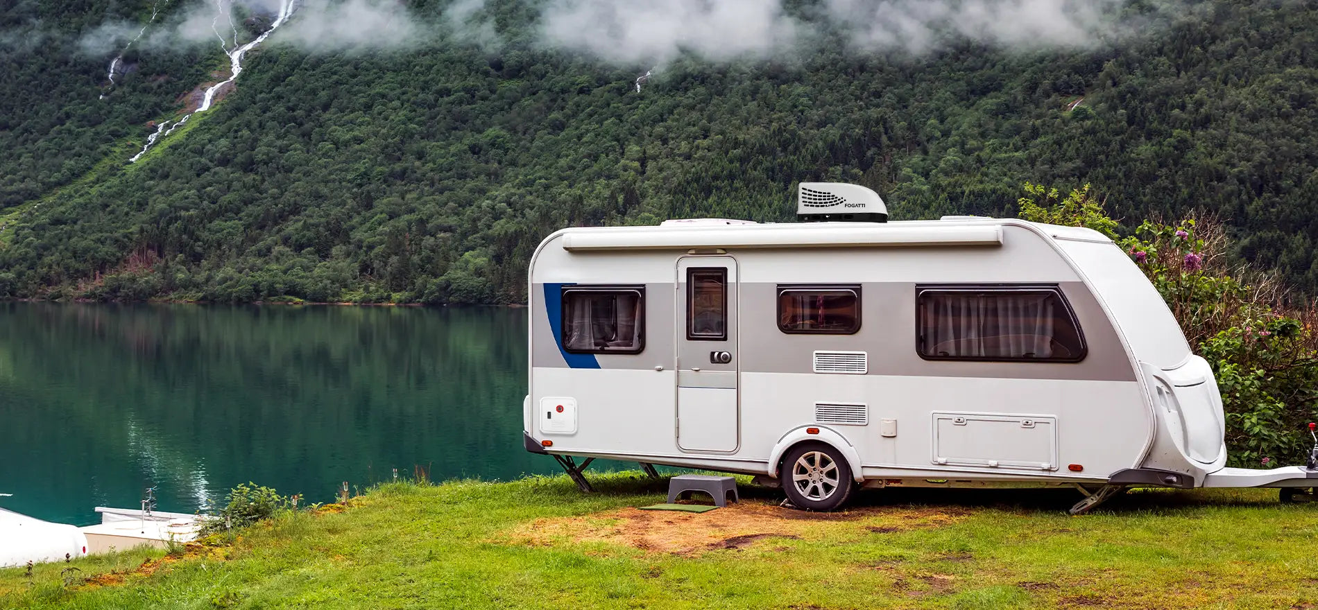 A white caravan with a Fogatti air conditioner on top is parked by a serene lake, surrounded by lush green mountains and clear water.