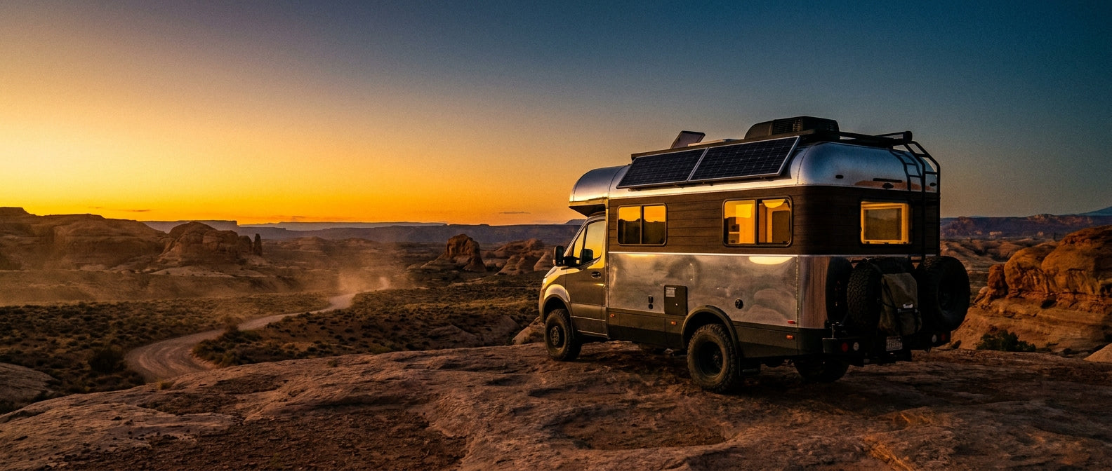 A modern RV parked on a desert plateau at golden hour with glowing interior lights against a dramatic amber and blue sky, suggesting off-grid comfort and adventure.